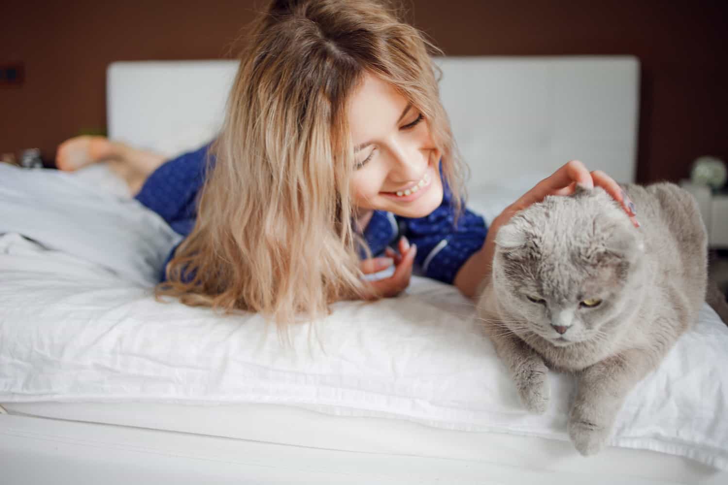 A woman lying on a bed, smiling and petting a gray cat