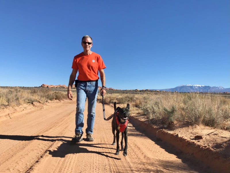 A man in an orange T-shirt walking with a smiling dog on Willow Flats Road in Arches National Park - Moab, UT