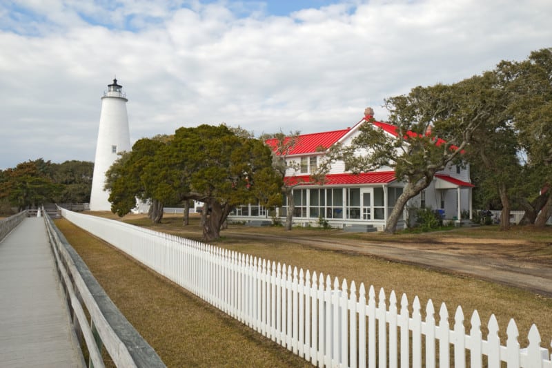 A white fence and a footpath lead past red-roofed guards to the lighthouse tower on Okracock Island on the outer coast of North Carolina, suitable for dogs.