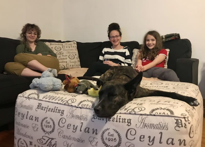A tiger dog is sleeping on an ottoman in front of two girls and a woman sitting on the couch